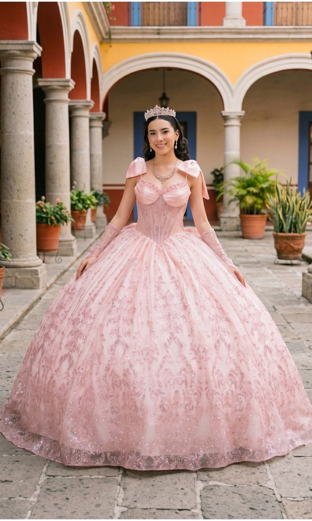A young woman in the Quinceanera Dress 8530JQ by Cinderella Couture, in a blush hue, stands in a courtyard adorned with stone arches and potted plants. She wears a sparkling tiara and delicate jewelry, while the beaded corset bodice of her gown shimmers softly, enhancing the elegant charm of her enchanting look.